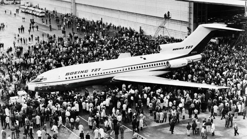 A black-and-white press photograph of the first Boeing 727 rolled out of its hangar, ringed by an enormous crowd of employees standing shoulder to shoulder around the aircraft and spilling back across the apron. The scale of the crowd is the point: the company the article is about was once a place people gathered to see what they had built.